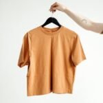 Minimalist shot of an orange t-shirt on a hanger held by a tattooed hand against a white background.